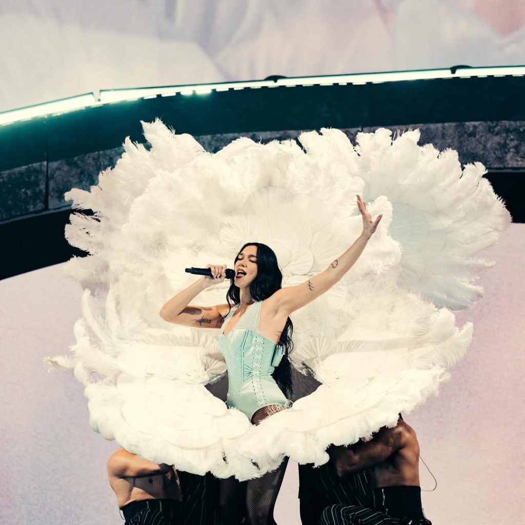 Dua Lipa cantando en el Radical Optimism Tour en Madrid con un impresionante vestuario celeste y abanicos de plumas blancos.