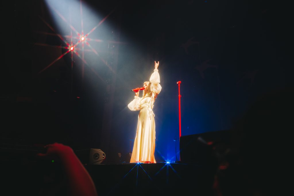 AURORA sobre el escenario levantando la mano con un gesto simbólico, micrófono rojo en mano y bañada por luces cálidas durante su concierto en Madrid